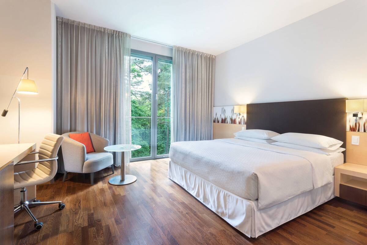 Ein modernes Hotelzimmer mit einem großen Bett, einem Schreibtisch, einem Sessel und Blick ins Freie durch das Fenster.