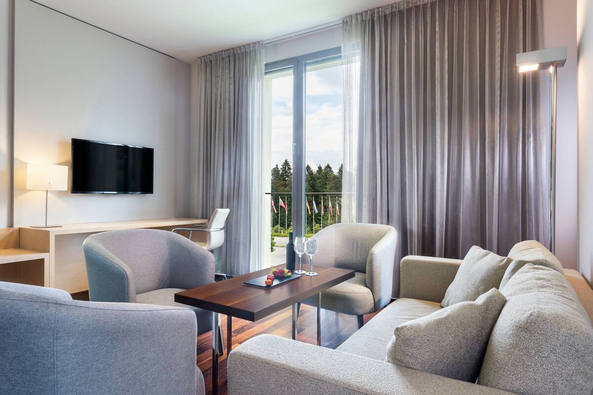 Ein modernes Hotelzimmer mit einem an der Wand befestigten Flachbildfernseher, einem Esstisch mit Stühlen, einer Sitzecke und einem Balkon mit Vorhängen.