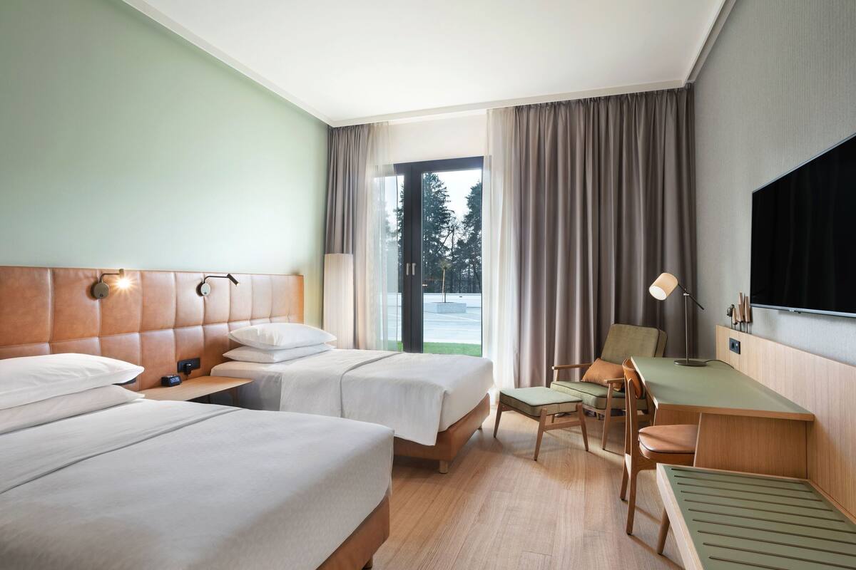 Ein Hotelzimmer mit zwei Betten, einem Schreibtisch, einem Sessel, einem Fernseher und einem großen Fenster mit Vorhängen.