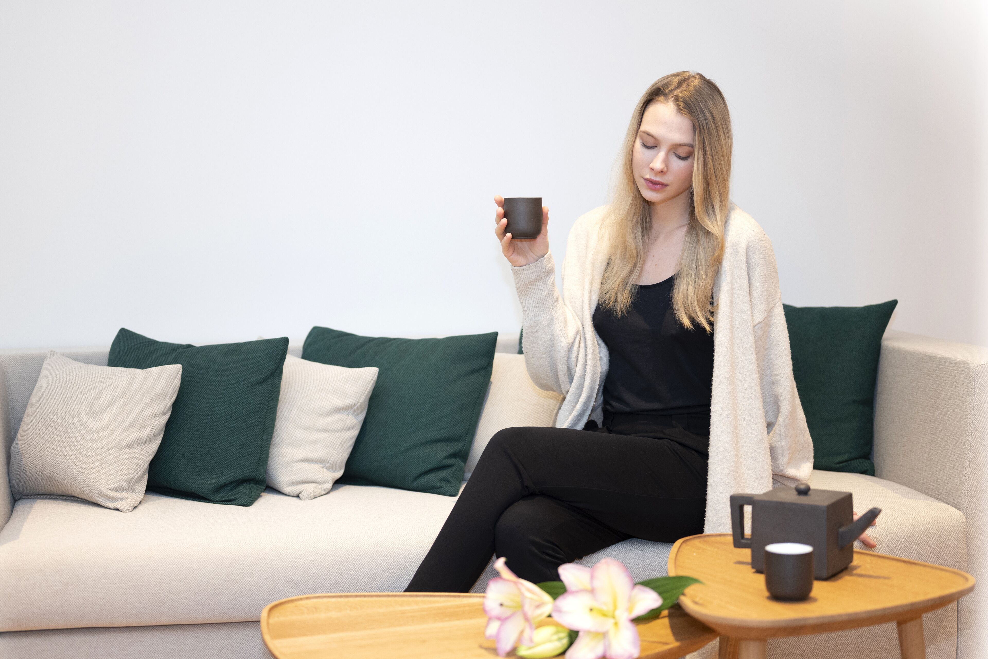 Eine Person sitzt auf einem Sofa, eine Tasse in der Hand, mit einer Teekanne und einem kleinen Tisch mit Blumen.