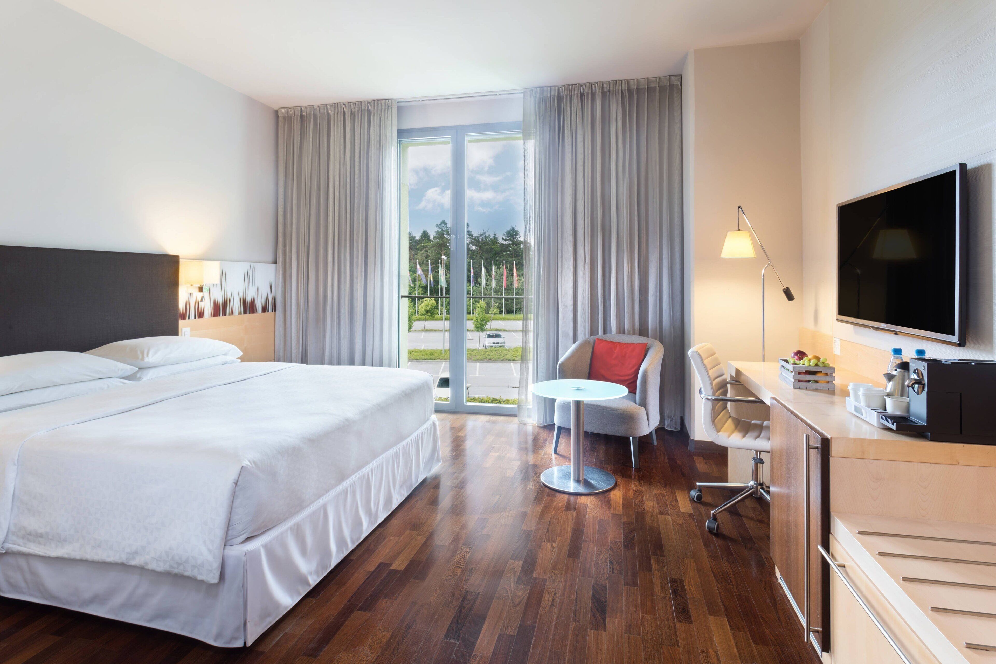 A hotel room with a large bed, a desk, a chair, a TV, and a balcony with a view of the outdoors.