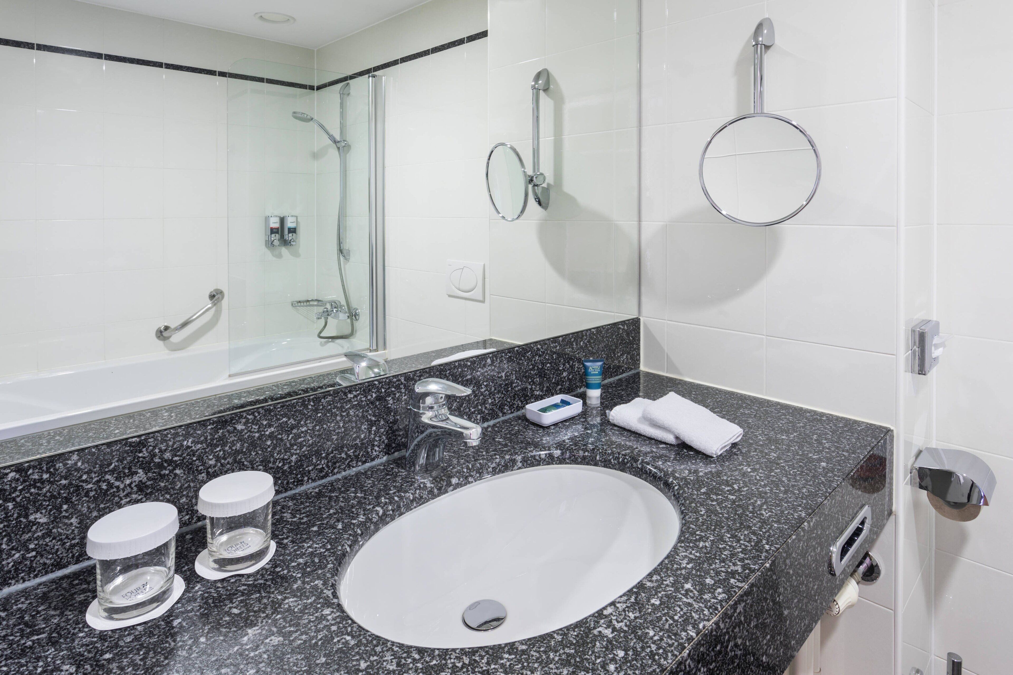 A bathroom with a granite countertop, a white sink, a mirror, a handheld showerhead, a towel rack, and a toilet paper holder.