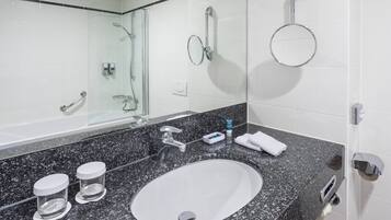 A bathroom with a granite countertop, a white sink, a mirror, a handheld showerhead, a towel rack, and a toilet paper holder.