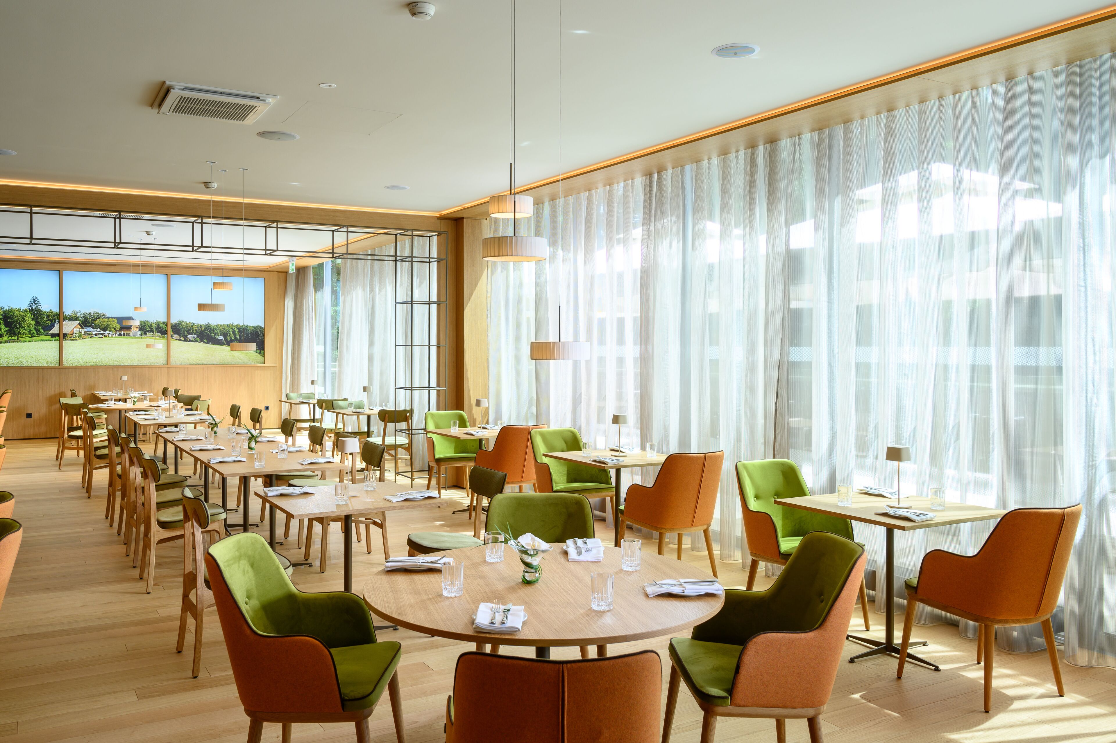 A spacious dining area with multiple tables and chairs, large windows with curtains, and a view of a green landscape.