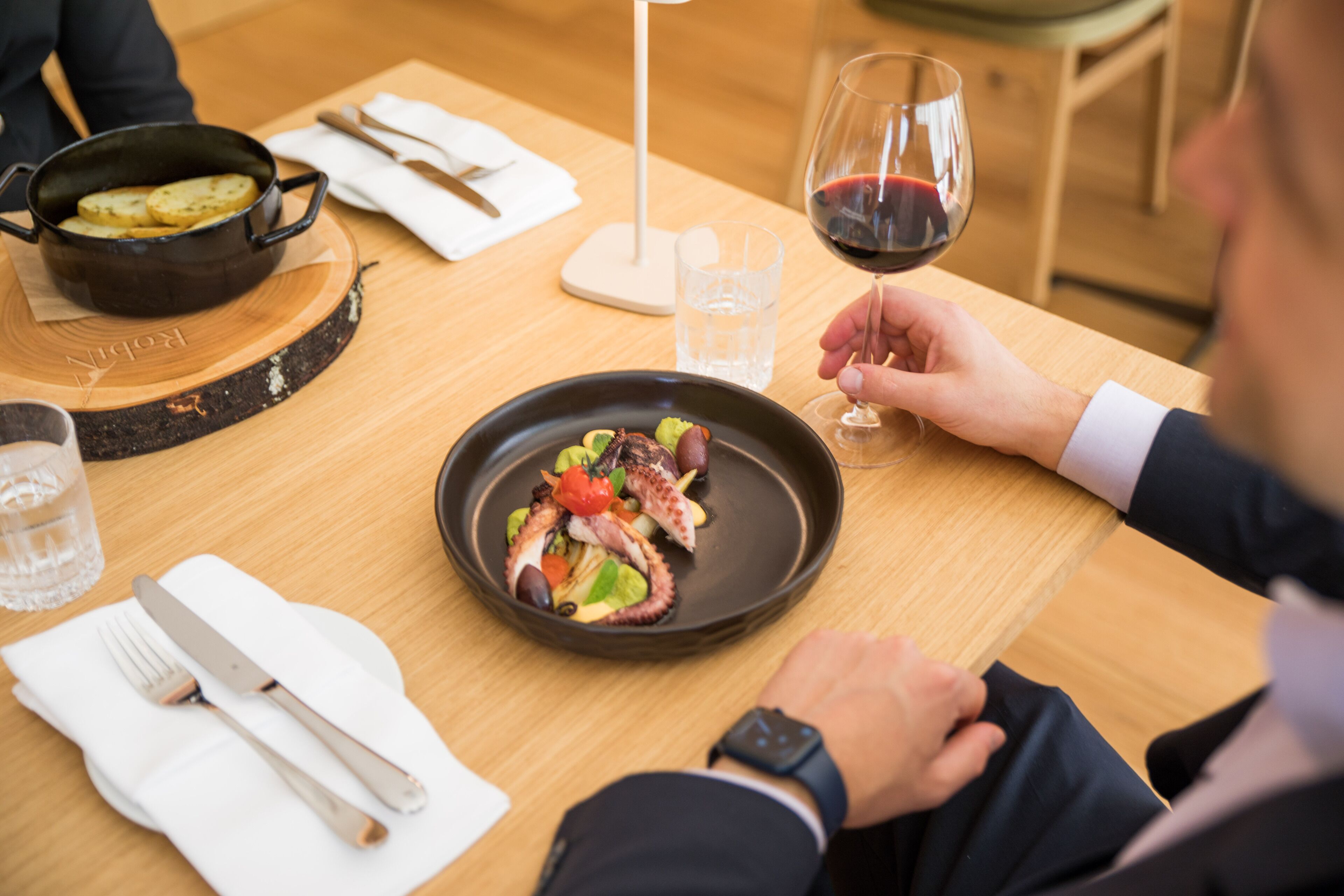A dining table with a plate of food, a wine glass, and a pot on a wooden board.