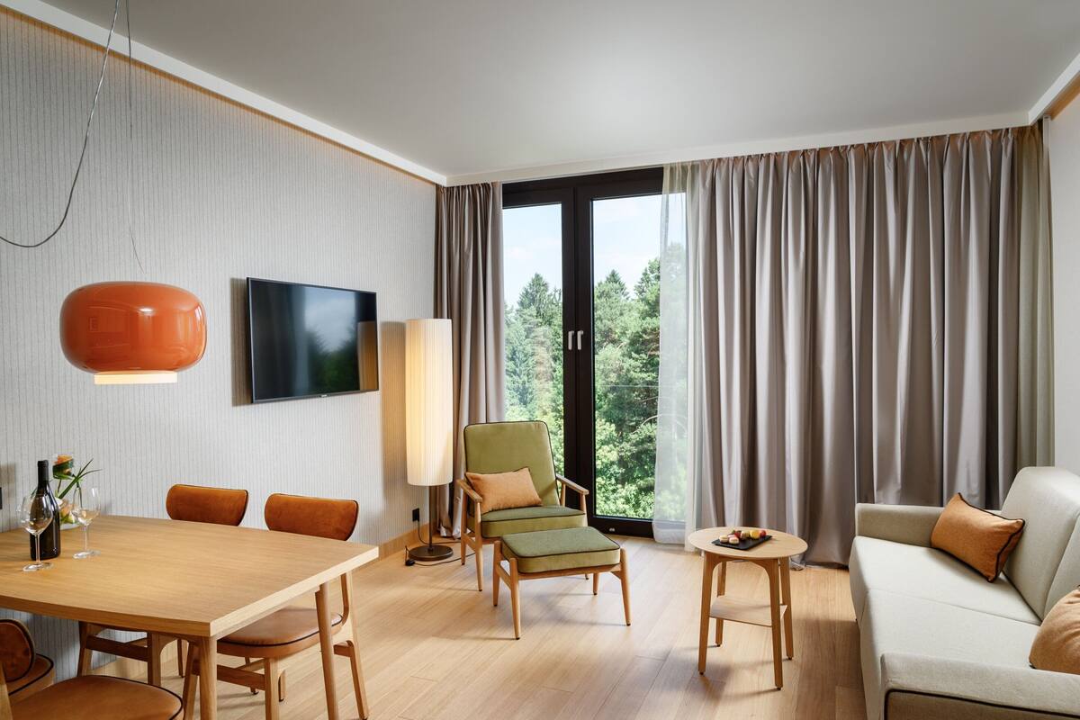 Ein modernes Hotelzimmer mit einem Holztisch, einer Couch, einem Sessel, einem Fernseher und einem großen Fenster mit Vorhängen.