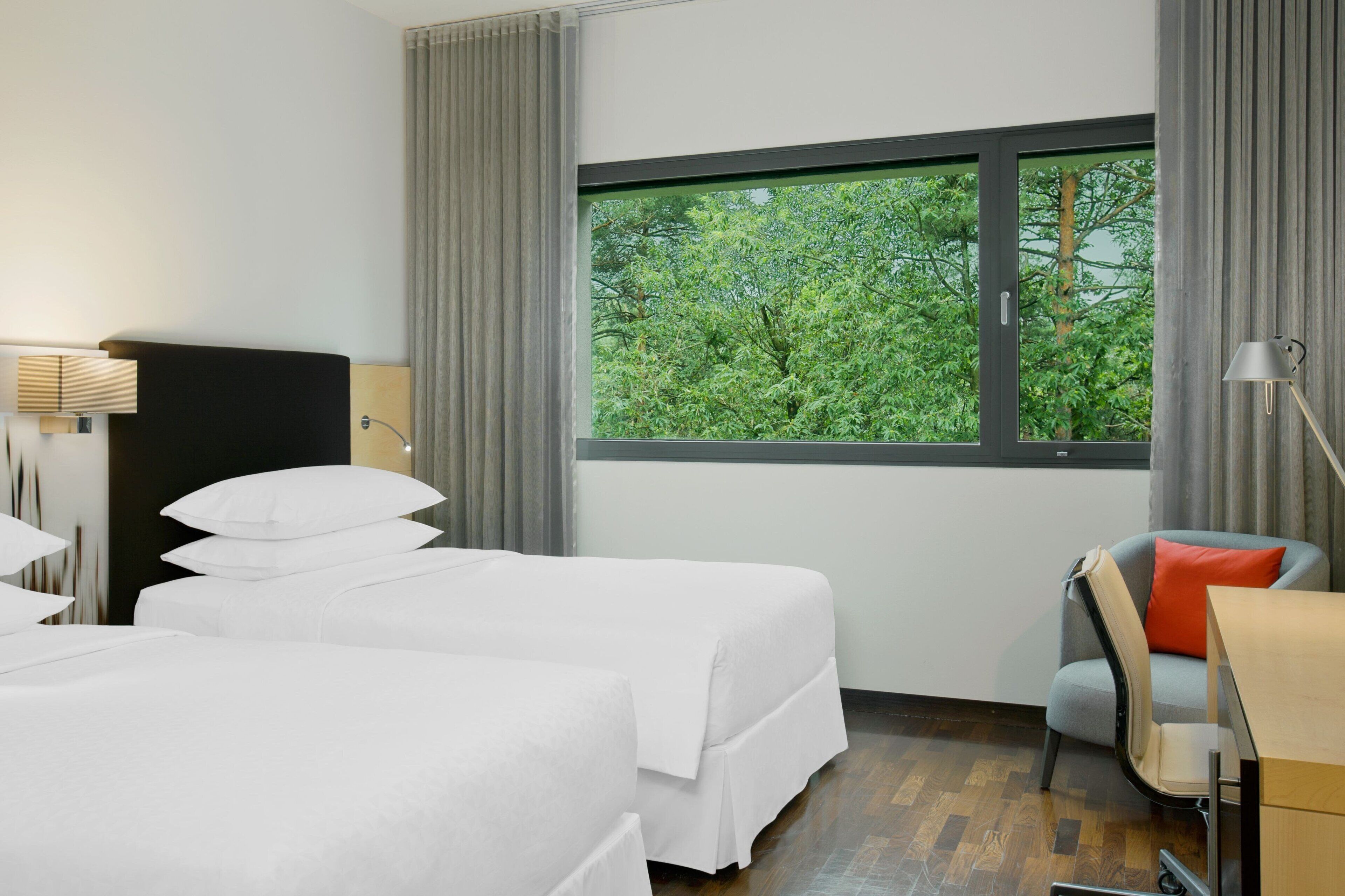 A hotel room with two beds, a desk, a chair, and a window with a view of greenery.