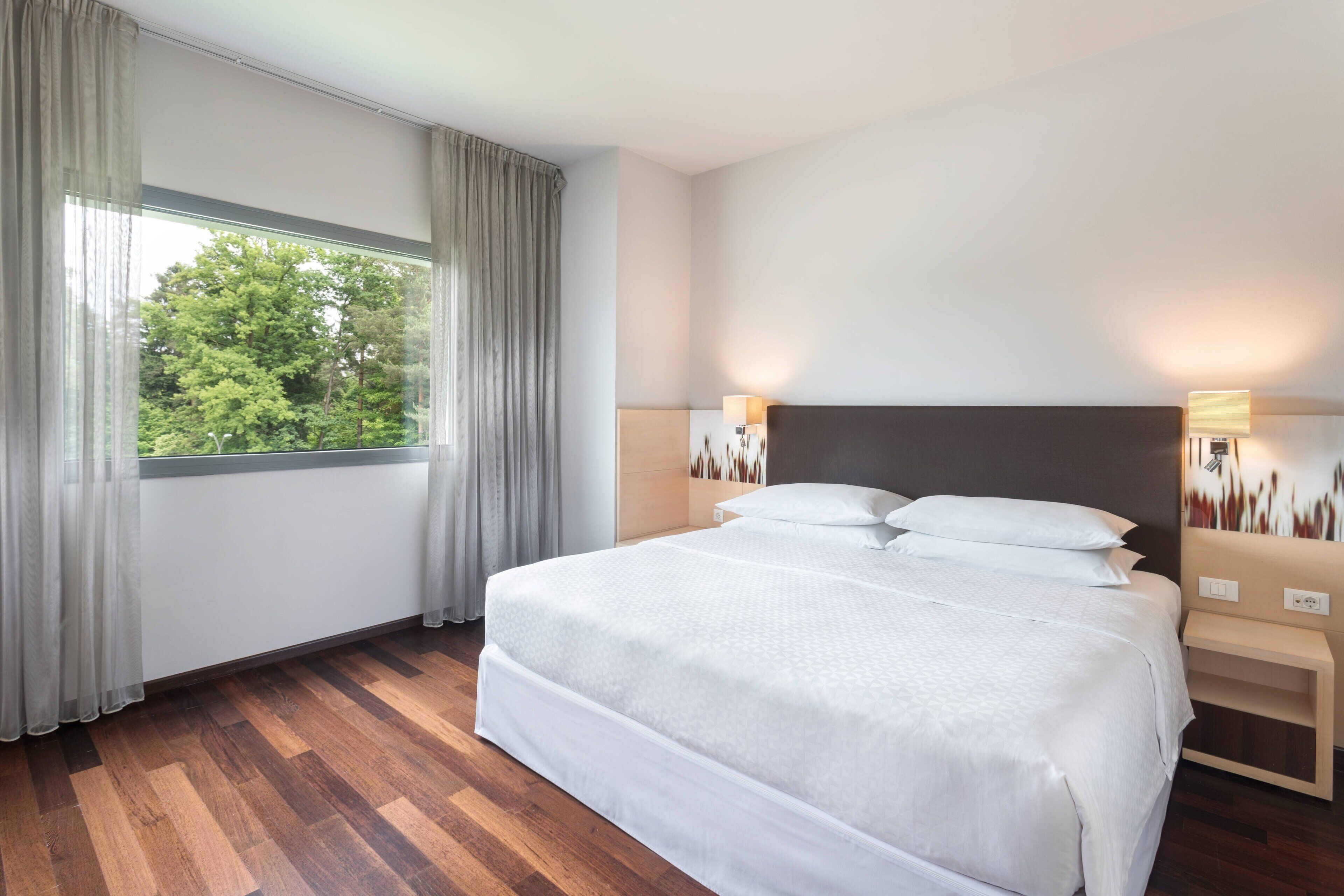 Ein modernes Hotelzimmer mit einem großen Bett, Holzböden und Blick auf Bäume durch das Fenster.