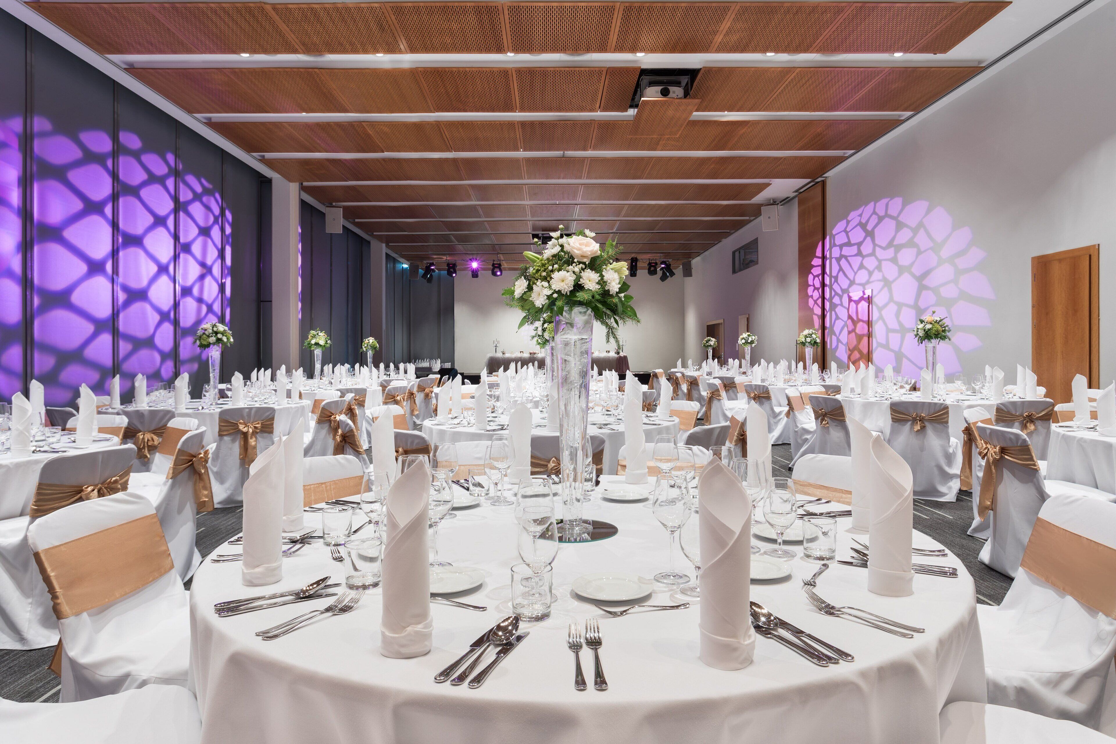 Un salón de banquetes con mesas redondas dispuestas para un evento formal, que incluye manteles blancos, cubertería y centros de mesa florales.