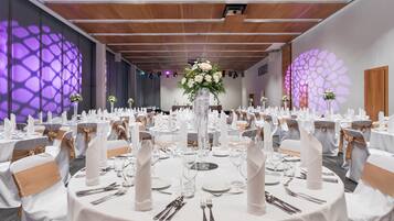 Un salón de banquetes con mesas redondas dispuestas para un evento formal, que incluye manteles blancos, cubertería y centros de mesa florales.