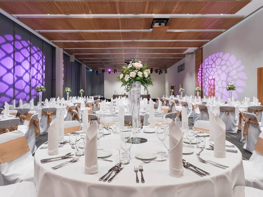 Un salón de banquetes con mesas redondas dispuestas para un evento formal, que incluye manteles blancos, cubertería y centros de mesa florales.