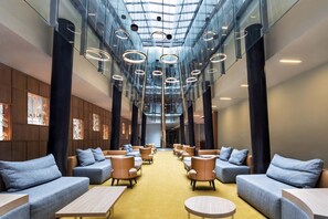 Un hall d’entrée moderne, avec des canapés et des tables disposés le long d’une allée centrale, de grandes fenêtres et un plafond haut orné de luminaires circulaires.