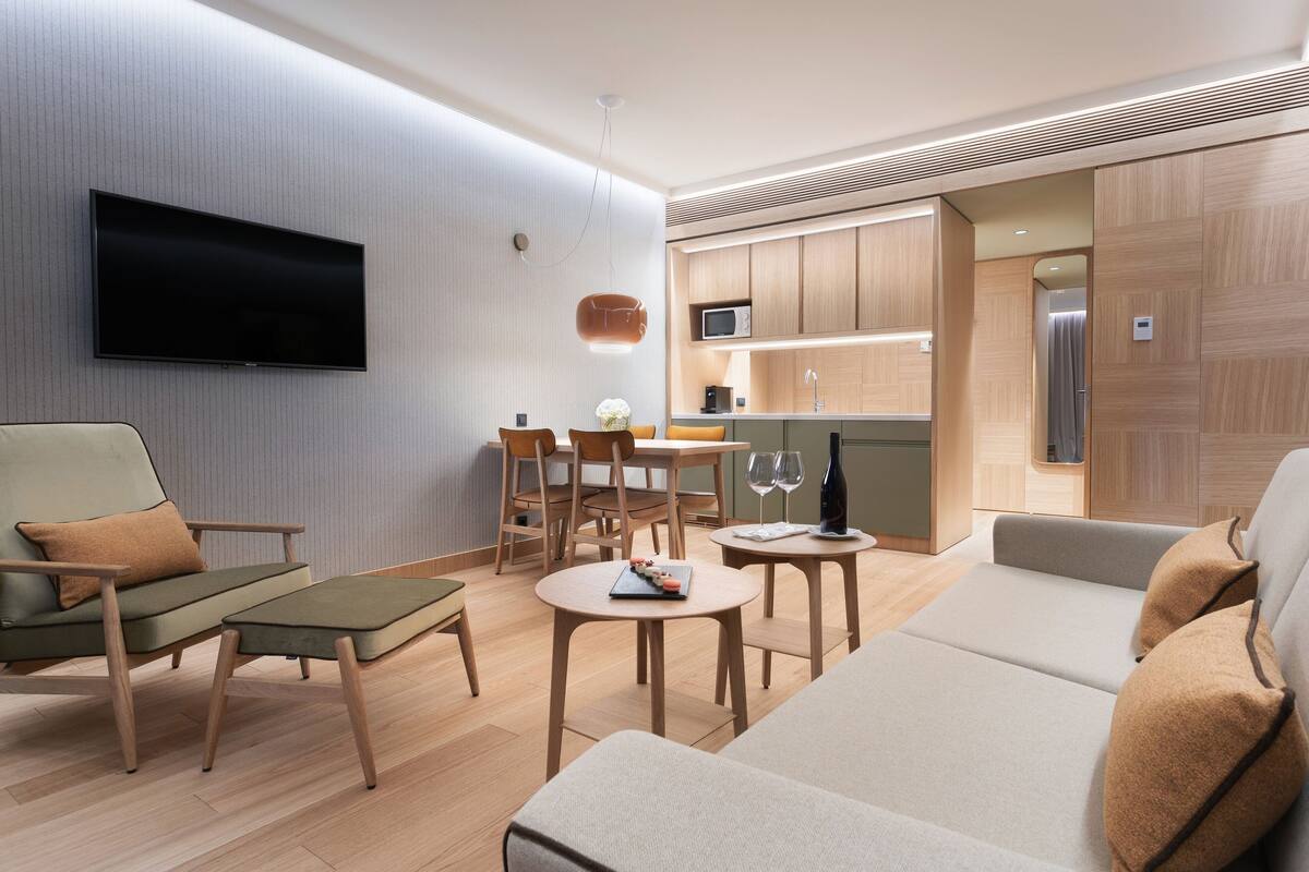 Ein modernes Wohnzimmer mit Flachbildfernseher, einem Essbereich mit Tisch und Stühlen sowie einer Küche mit Holzschränken und einer Mikrowelle.