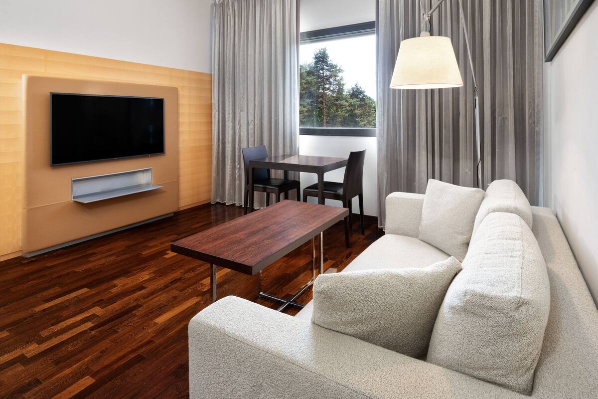 Ein modernes Wohnzimmer mit Flachbildfernseher, Holzboden, einer Couch, einem Esstisch und einem Fenster mit Vorhängen.