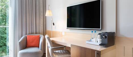 Ein modernes Hotelzimmer mit Schreibtisch aus Holz, zwei Stühlen, einem Flachbildfernseher, einer Lampe und einer Kaffeemaschine.