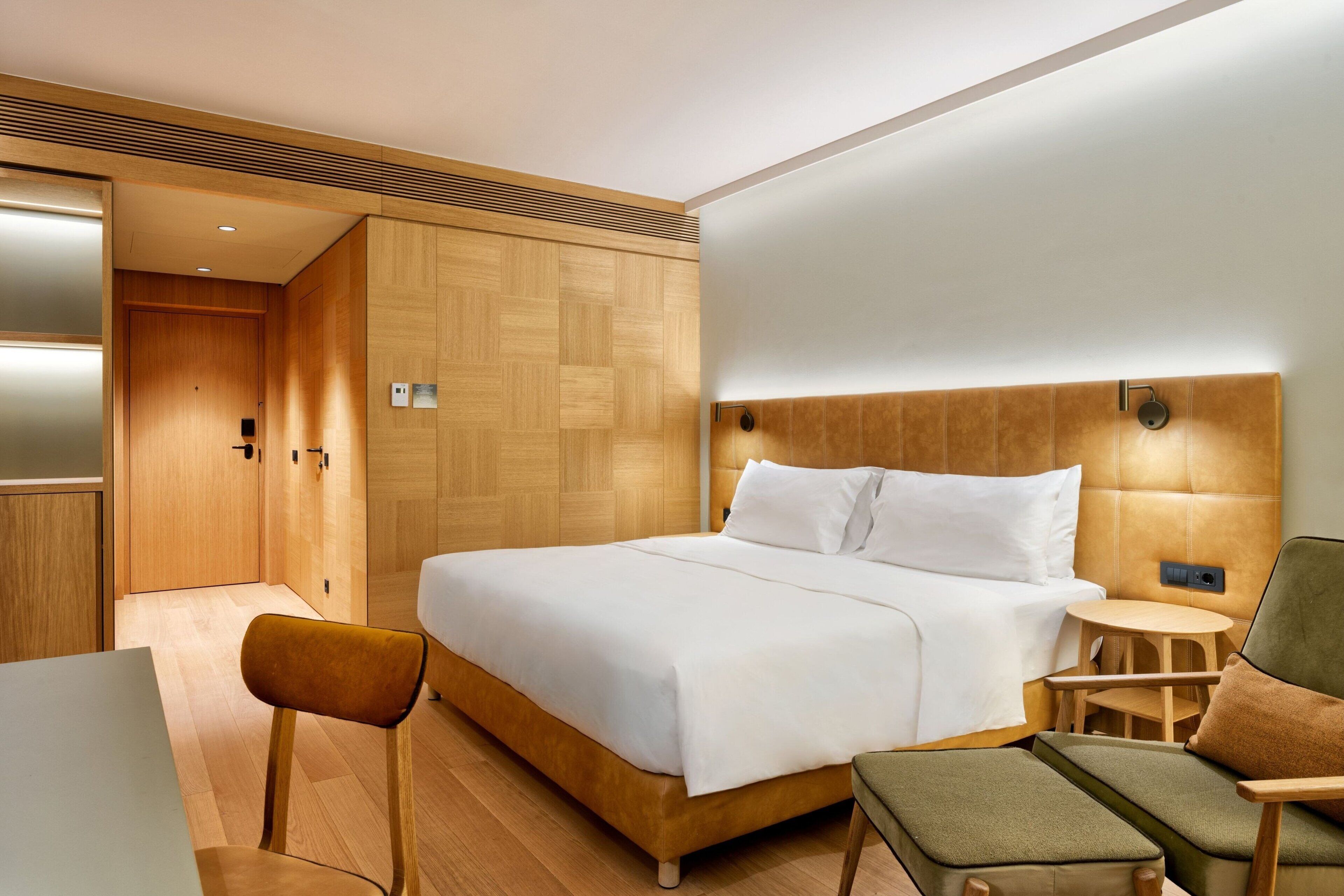 A modern hotel room with a large bed, a wooden headboard, a small round table, and a green armchair.