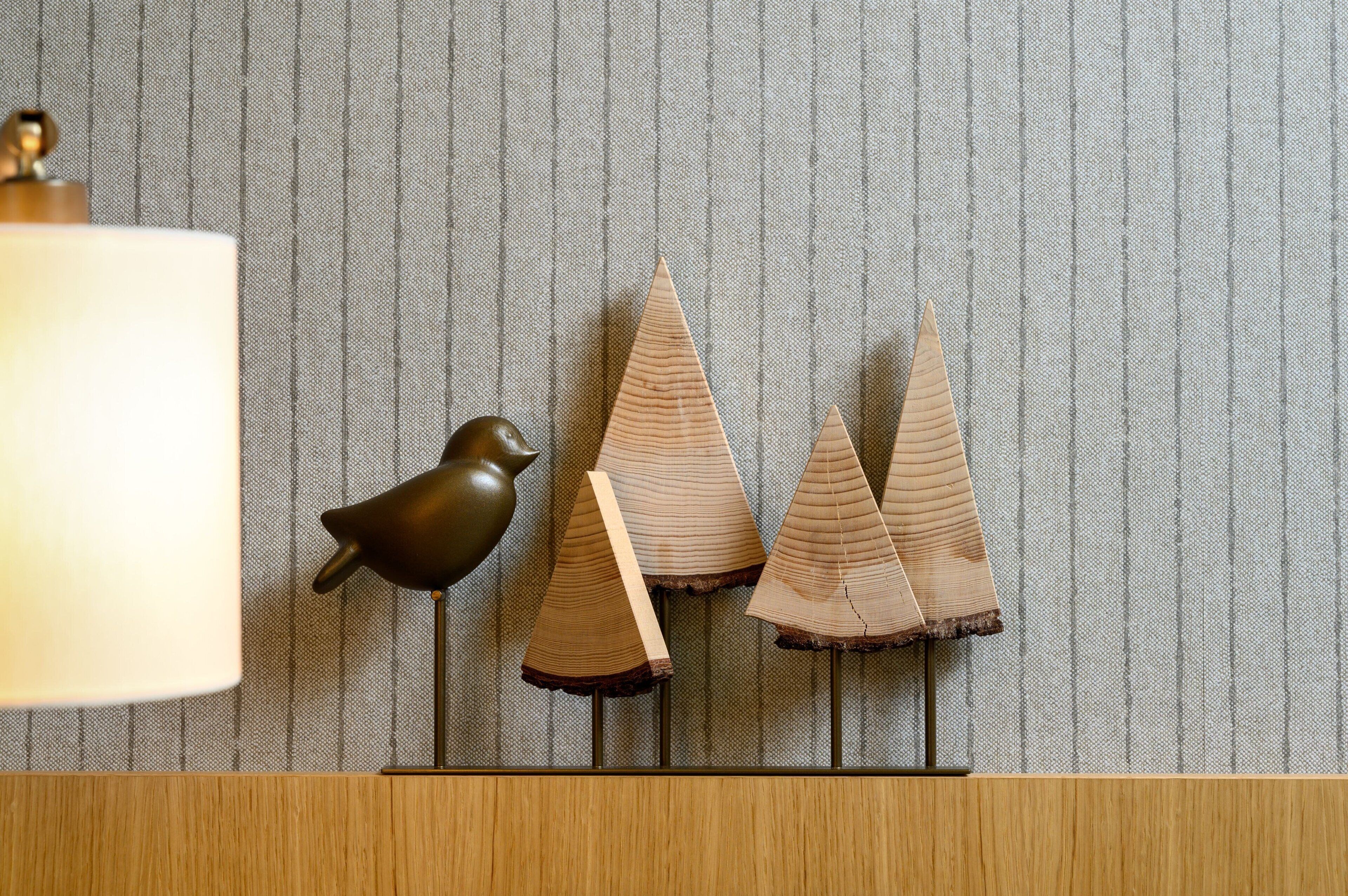 Una lámpara, una figurita de pájaro y tres objetos de madera con forma de cono sobre un pedestal frente a una pared texturizada.