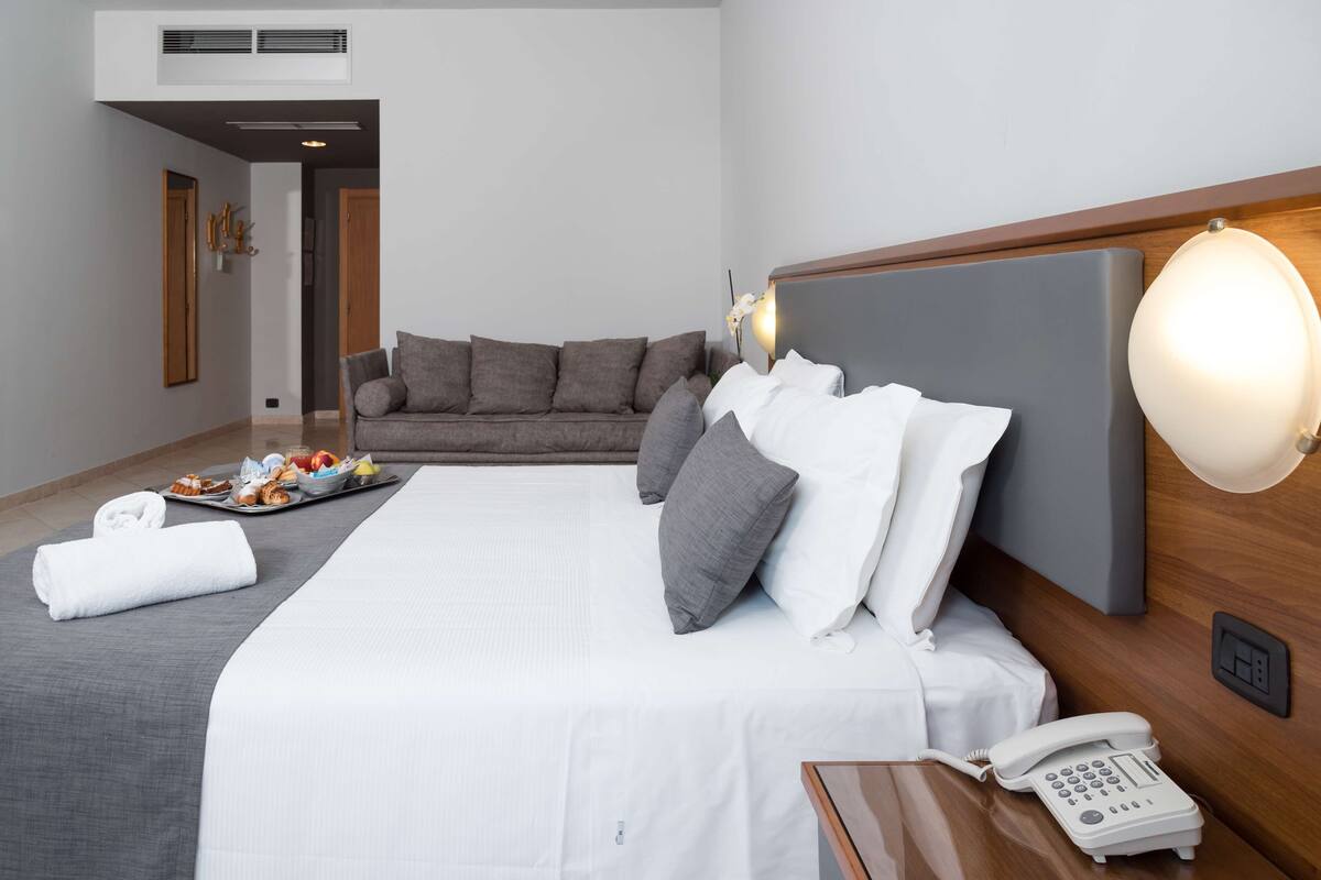 A hotel room with a large bed, a bedside table with a phone, a sofa, and a tray of breakfast items.