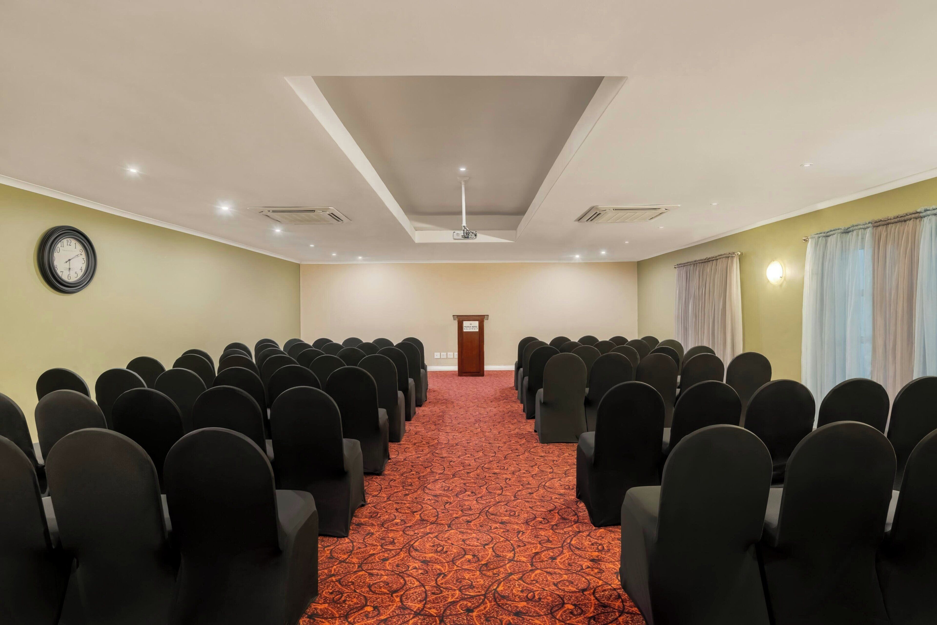 A conference room with rows of chairs, a carpeted floor, a podium, and a clock on the wall.
