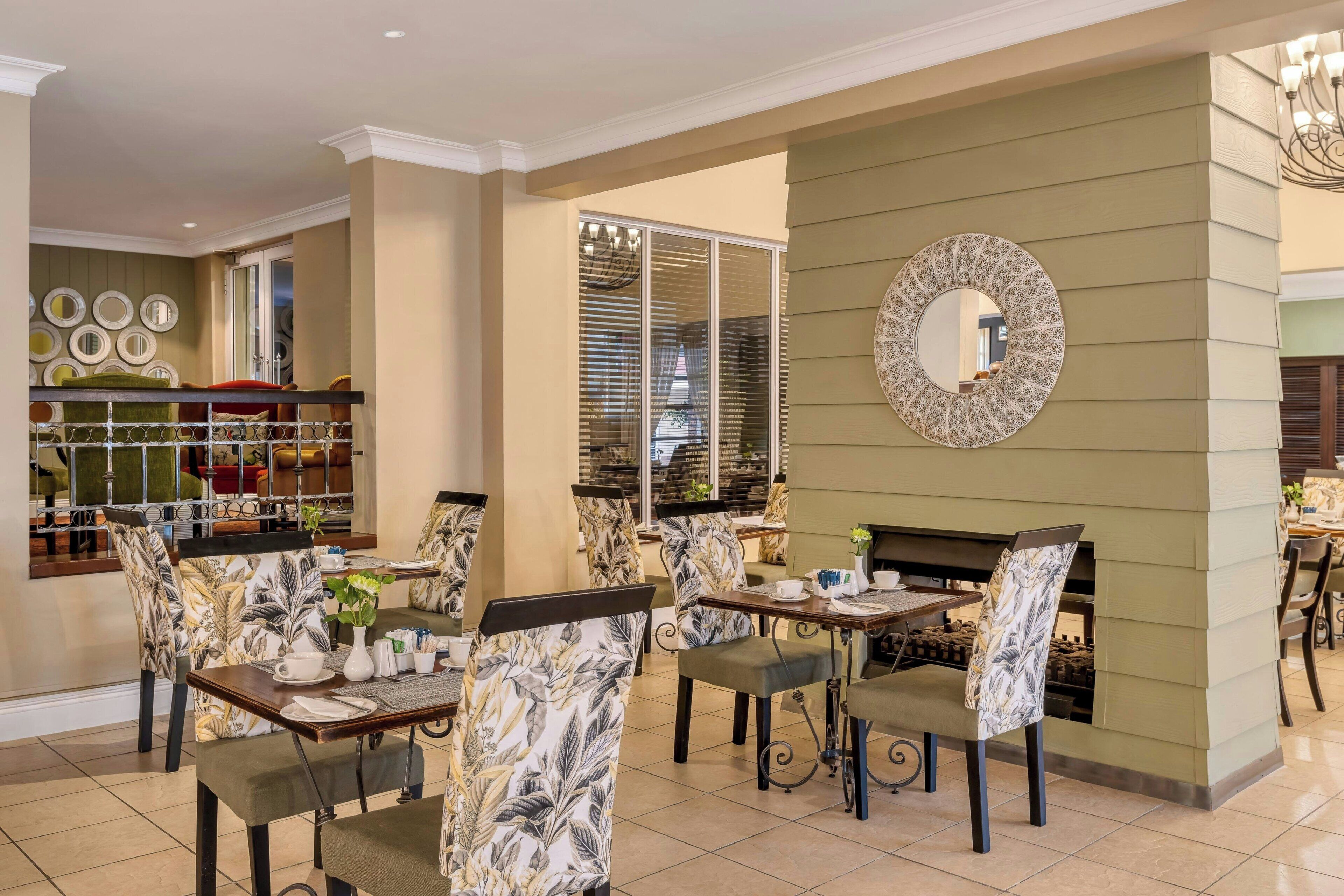 A dining area with patterned chairs, a round mirror, and a view of the kitchen through a glass door.