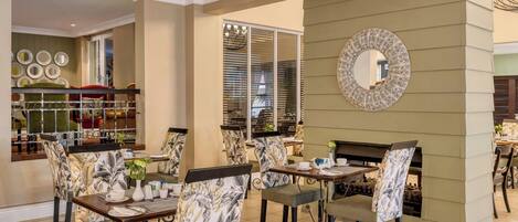 A dining area with patterned chairs, a round mirror, and a view of the kitchen through a glass door.