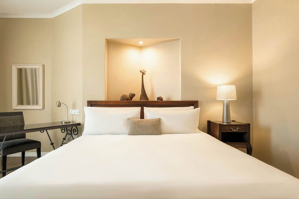 Una habitación de hotel con cama, escritorio, silla, mesita de noche con lámpara y espejo montado en la pared.