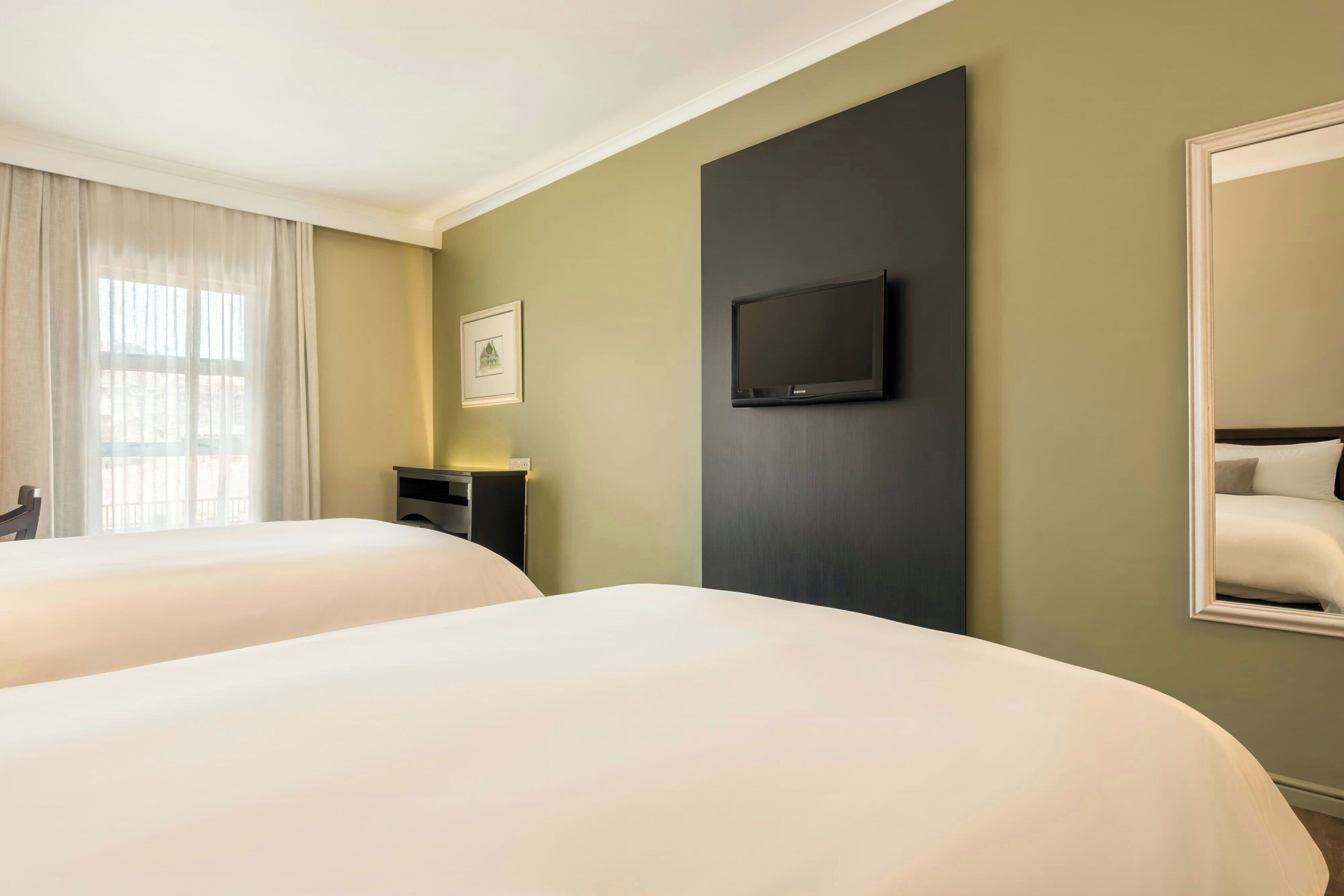 A hotel room with two beds, a flat-screen TV mounted on the wall, a bedside table, and a window with curtains.