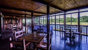 An indoor dining area with wooden tables and chairs, large windows offering a view of greenery outside. - Imvubu Lodge (Richards Bay)