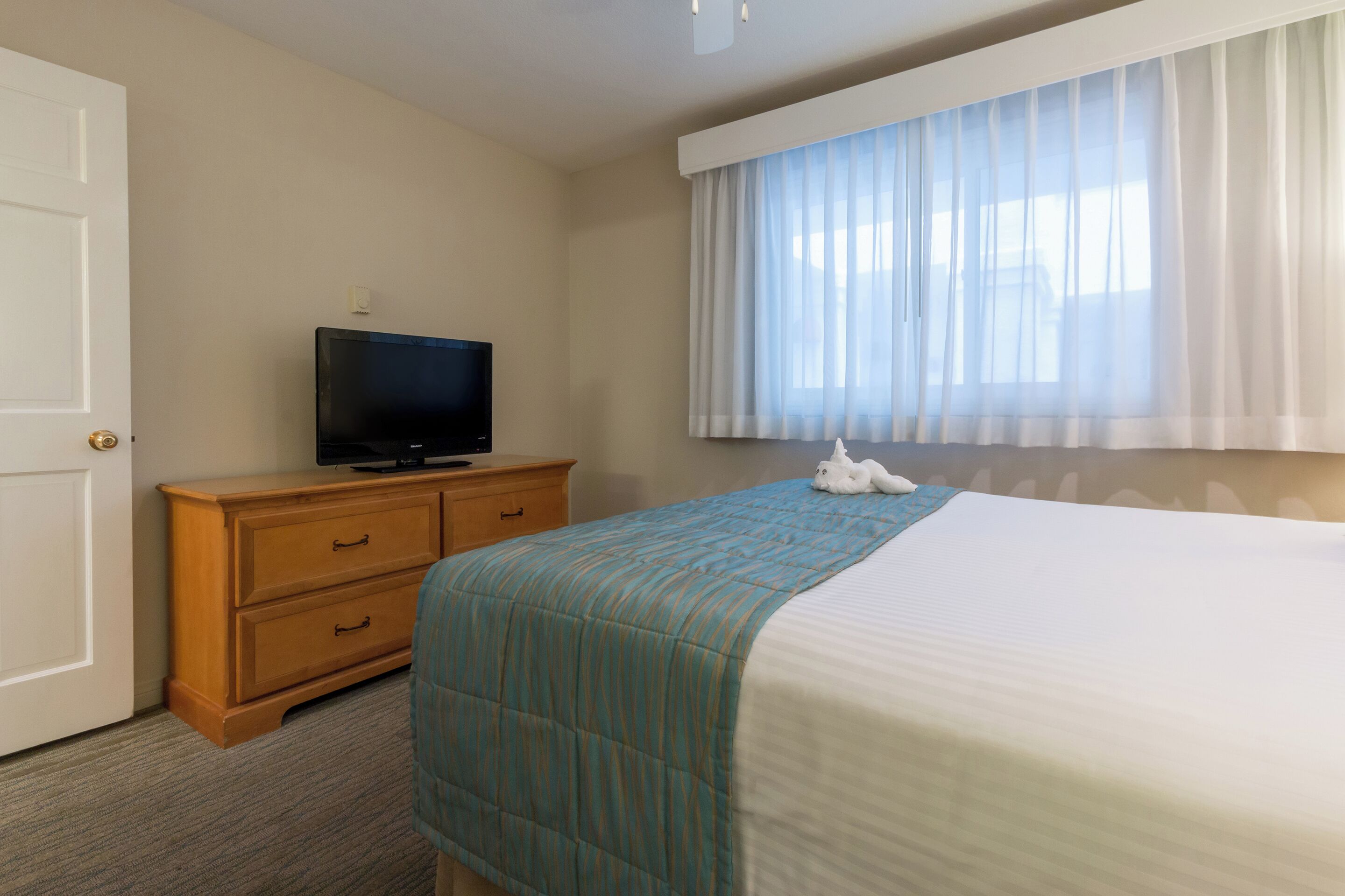A hotel room with a bed, a TV on a wooden dresser, and a window with sheer curtains.
