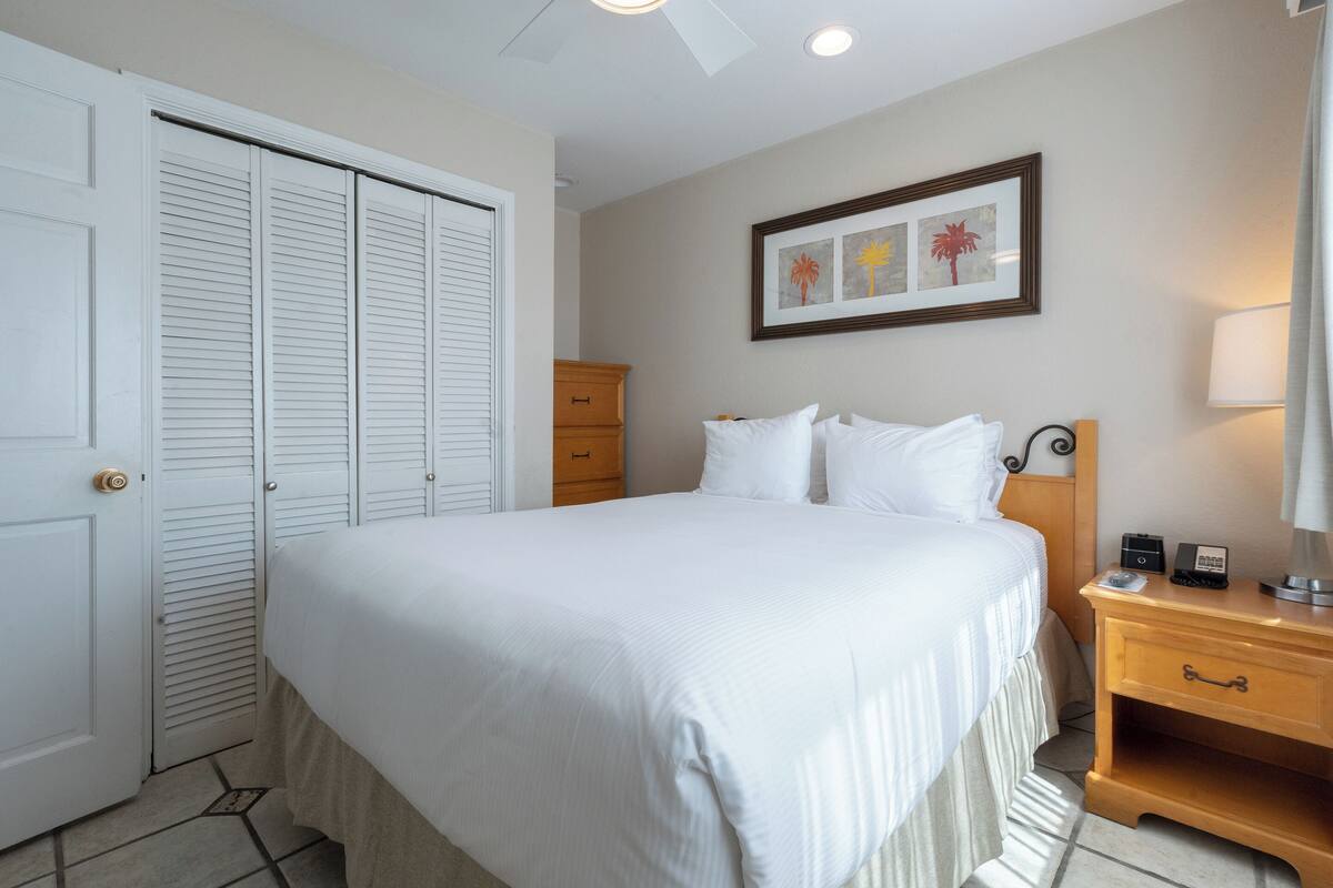 A neatly arranged bedroom with a large bed, a wooden nightstand, a dresser, and a framed picture on the wall.