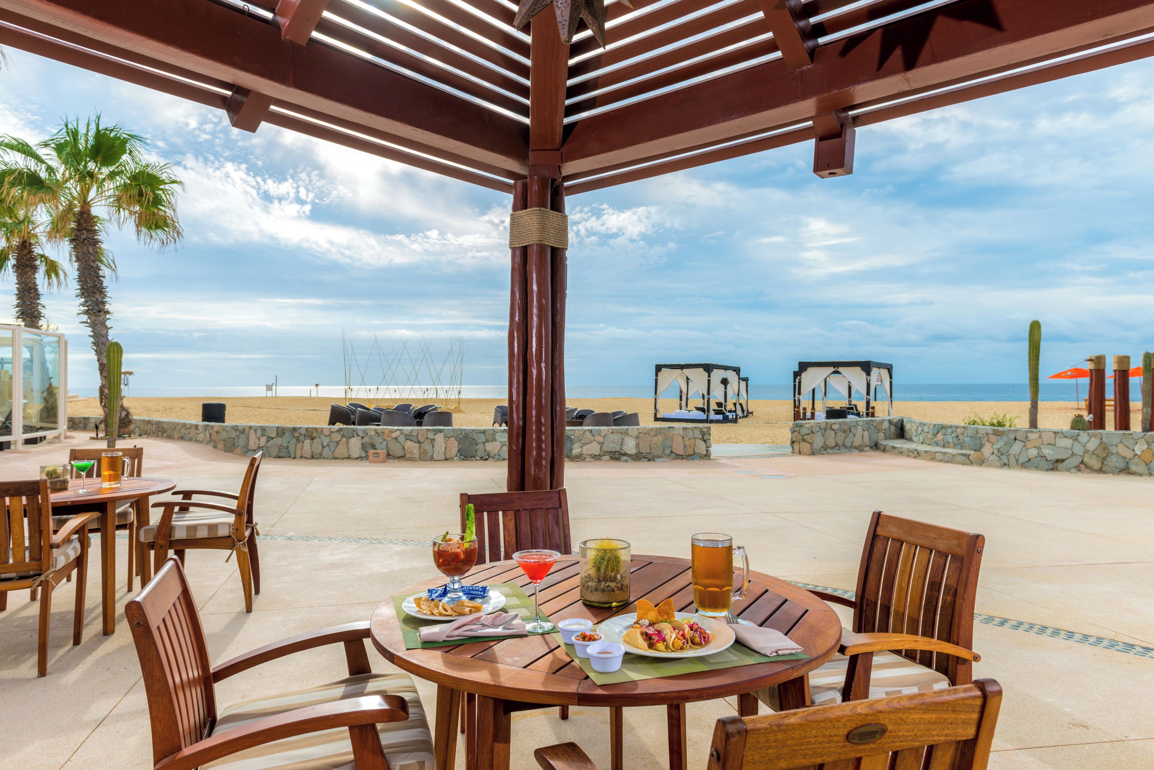 An outdoor dining area with a view of the beach and ocean, featuring wooden tables and chairs, a variety of drinks, and a stone wall.