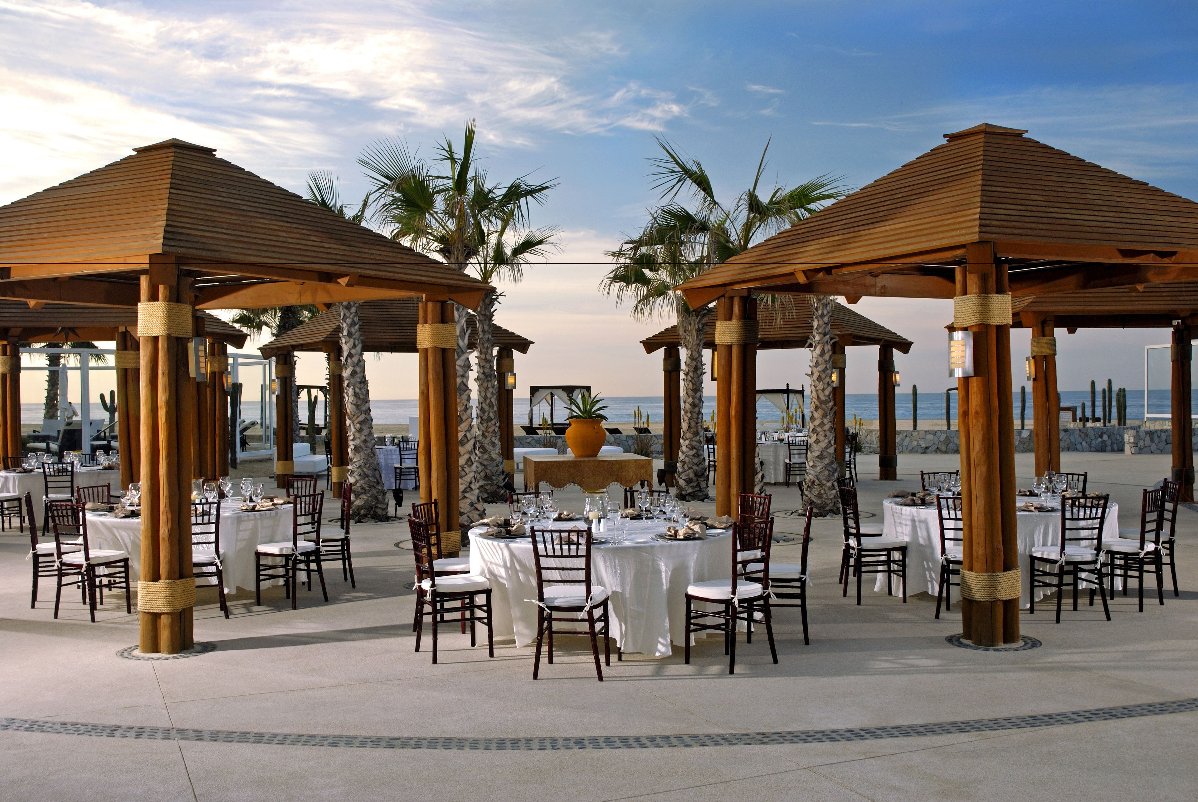Een restaurant aan het strand met houten prieelvormige overkappingen, witte tafellakens en stoelen klaar voor dineren.