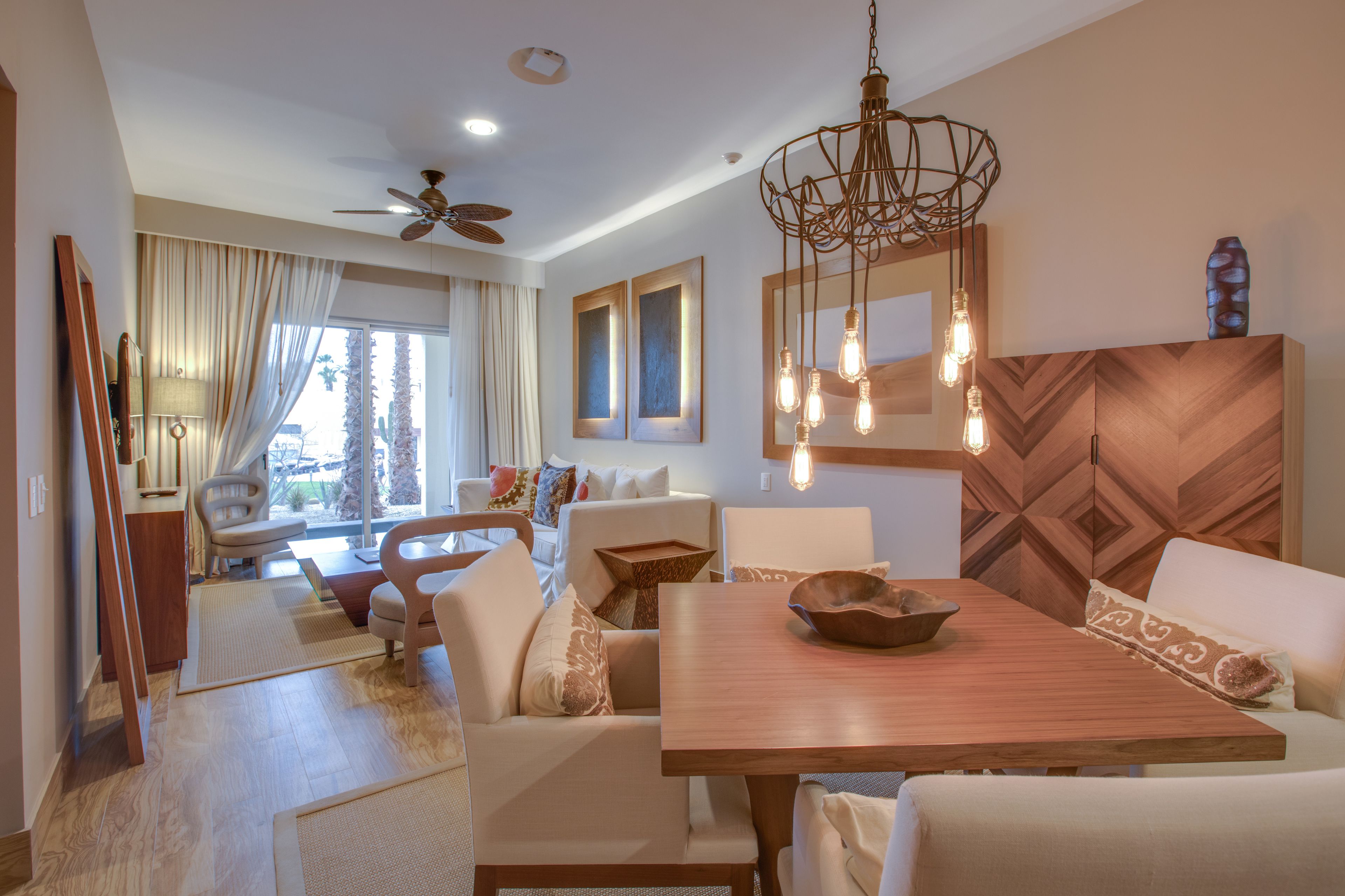 A spacious living room with a wooden dining table, comfortable seating, a ceiling fan, and a large window with curtains.