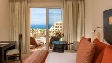 Ein Hotelzimmer mit Bett, Schreibtisch, Stuhl, einem kleinen Tisch mit Blumenvase und Blick auf das Meer und die Gebäude.