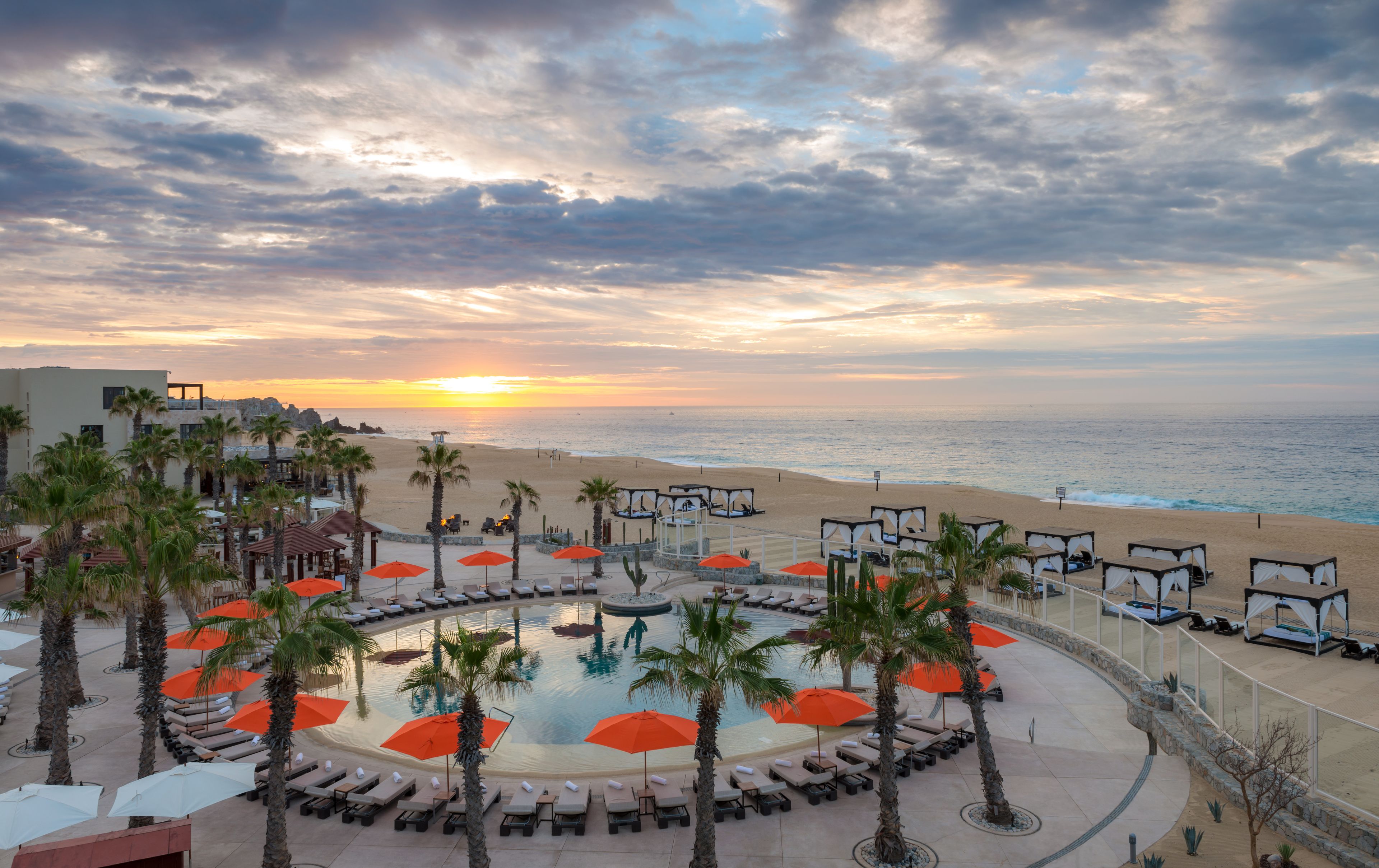 Ein Resort mit Pool, Liegestühlen und Sonnenschirmen mit Blick auf einen Strand und das Meer bei Sonnenuntergang.
