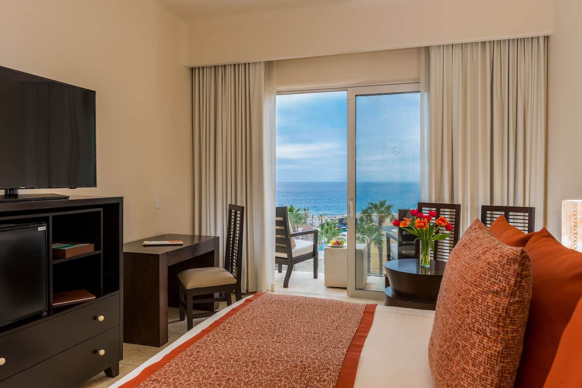 Une chambre d’hôtel avec un lit, un bureau, une télévision, un balcon donnant sur l’océan et une table décorée de fleurs.