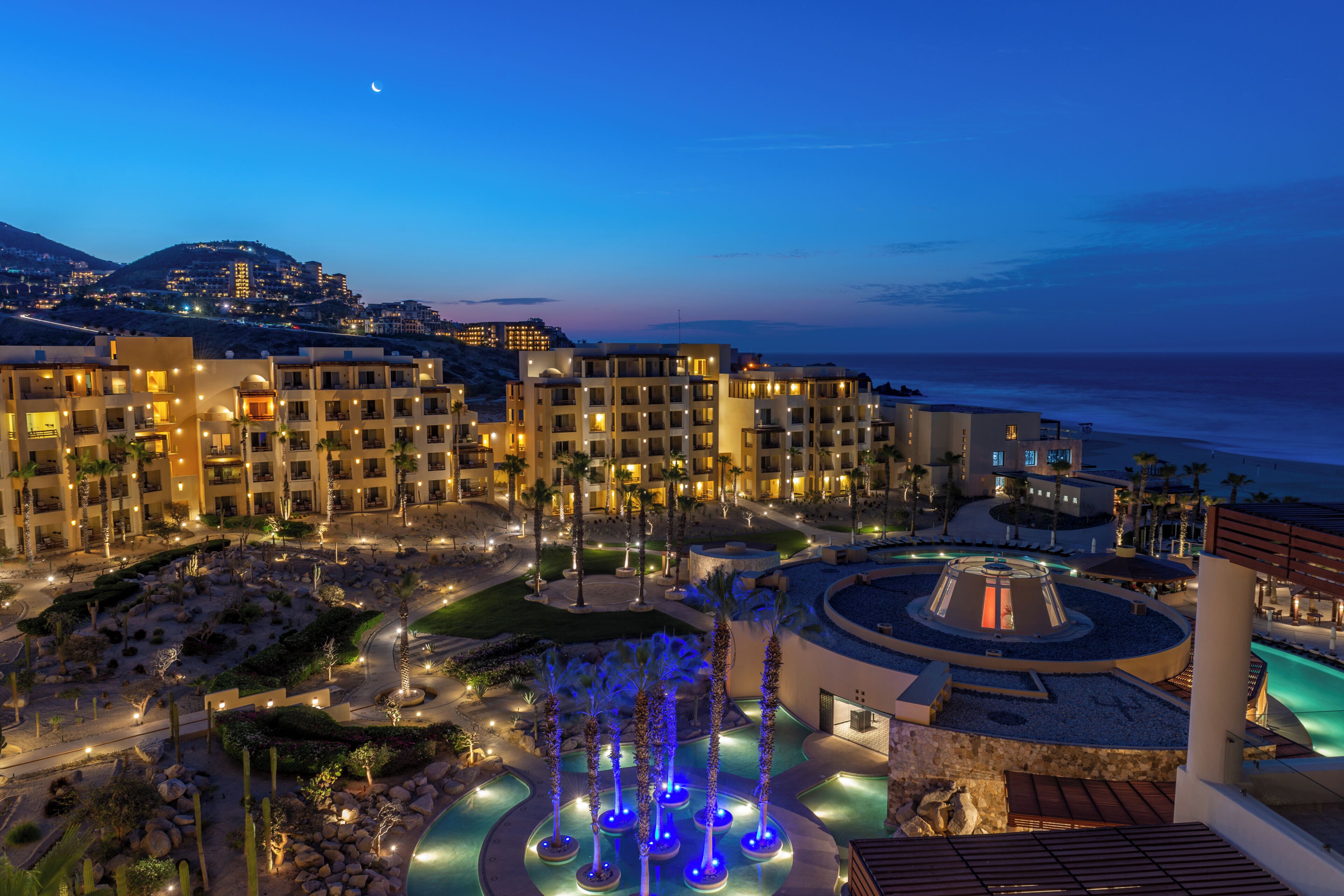 Un resort con varias edificaciones, un área de piscina ajardinada con una fuente y vista al océano.