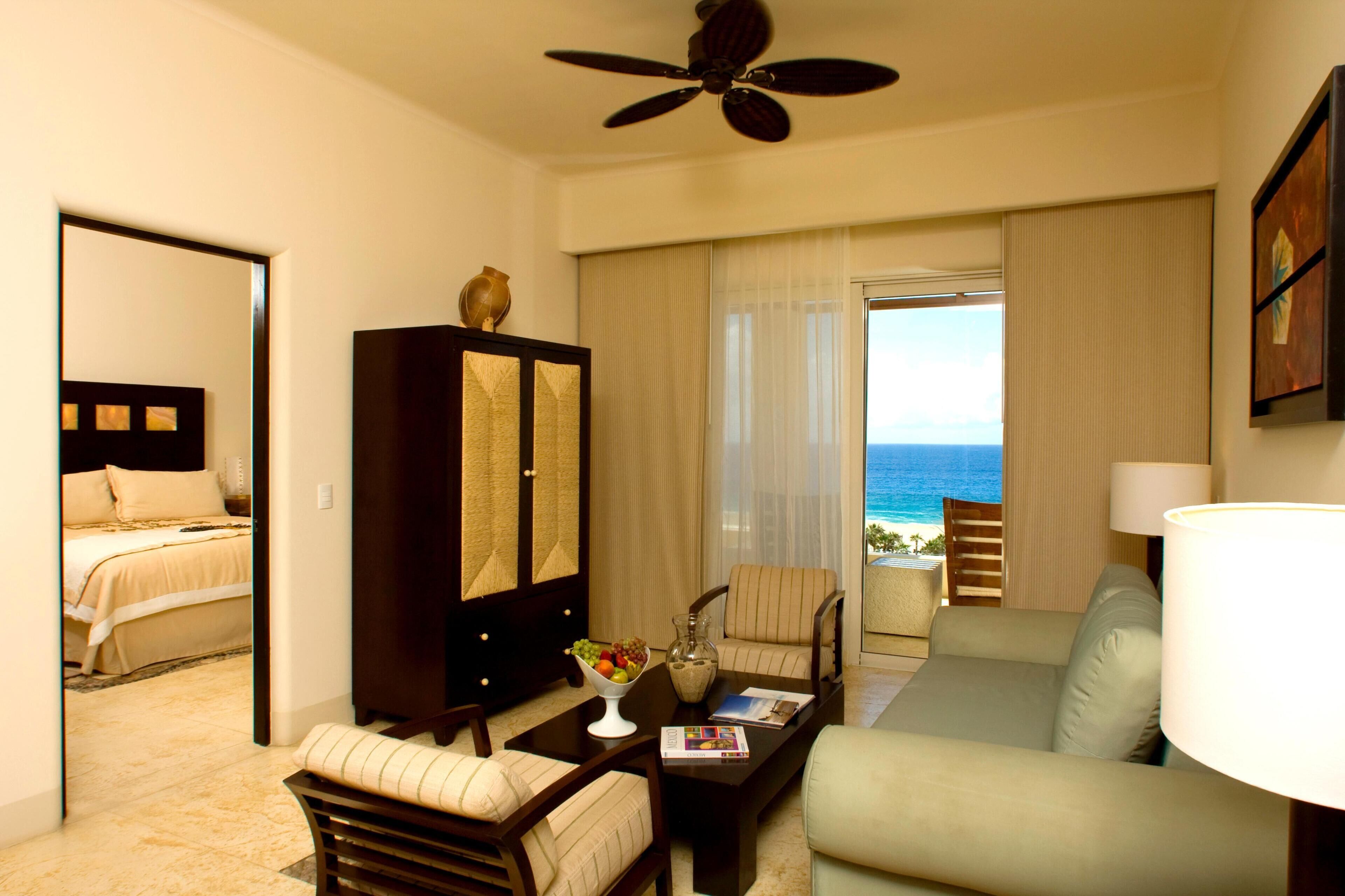 A hotel room with a bed, a seating area with a sofa and chairs, a coffee table, a lamp, and a view of the ocean.
