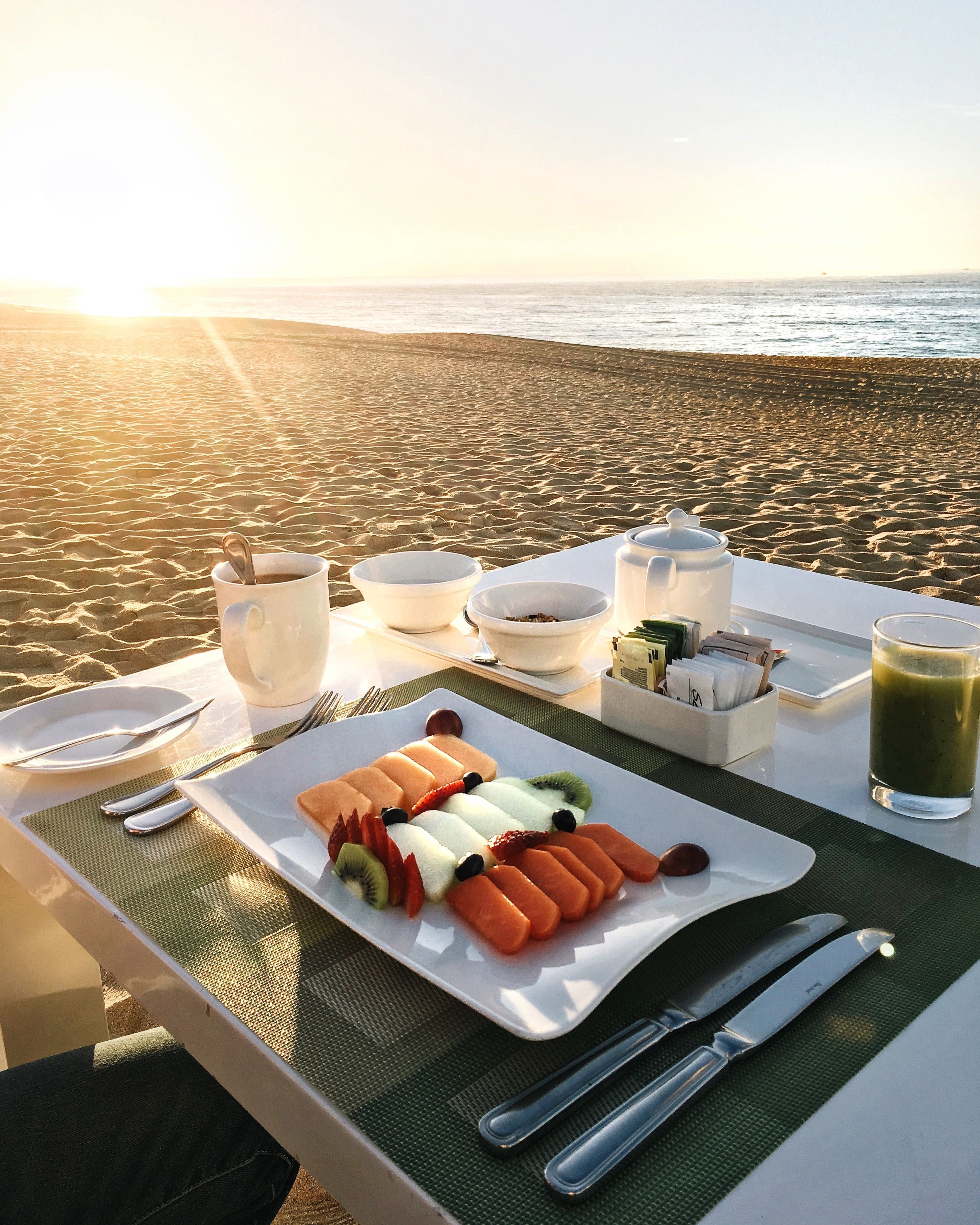 Un petit-déjeuner est préparé sur la plage, avec une table, des assiettes, des couverts et un verre de jus vert.