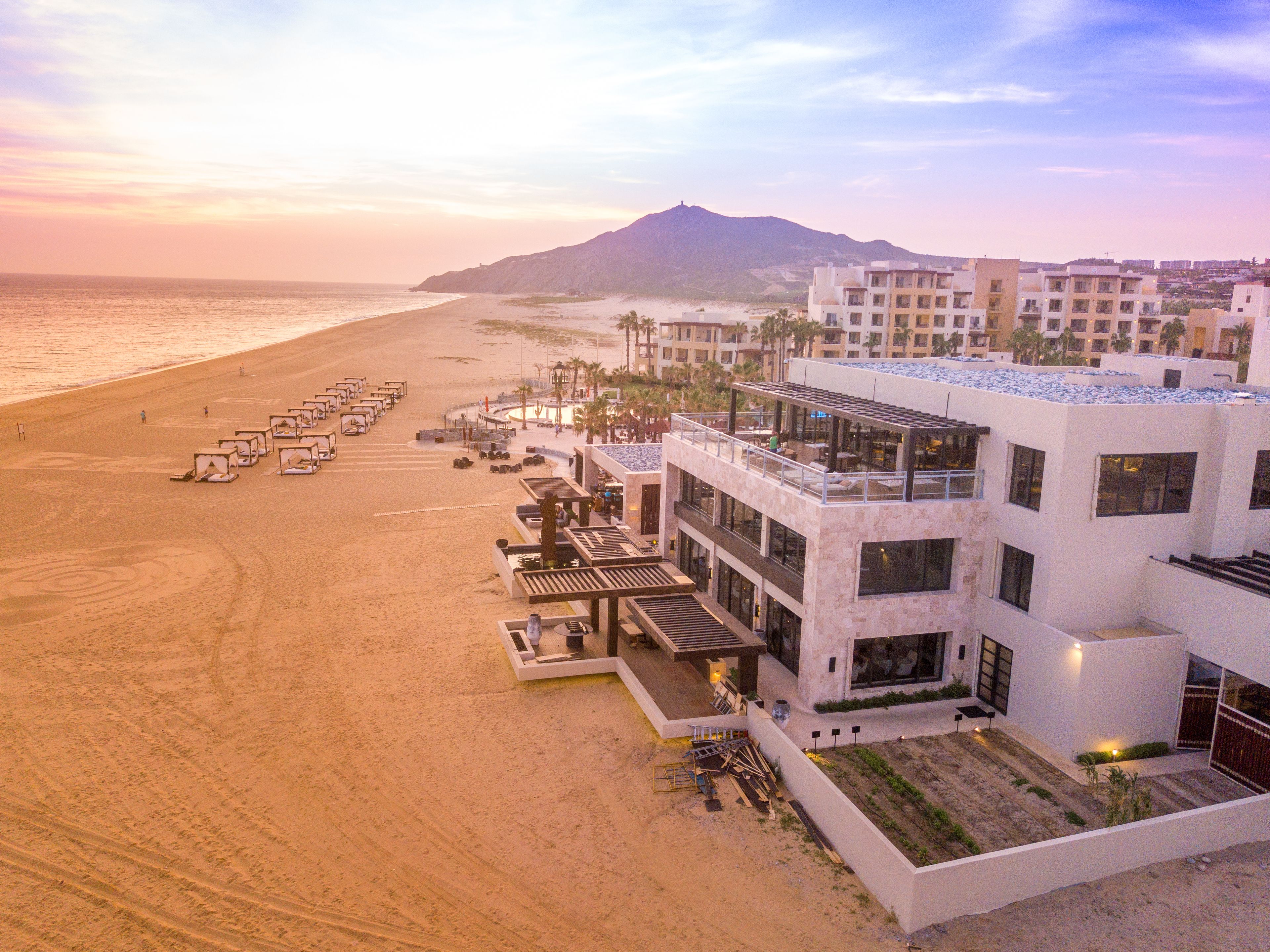 Un hotel frente a la playa con varias construcciones, una playa de arena y una montaña al fondo.