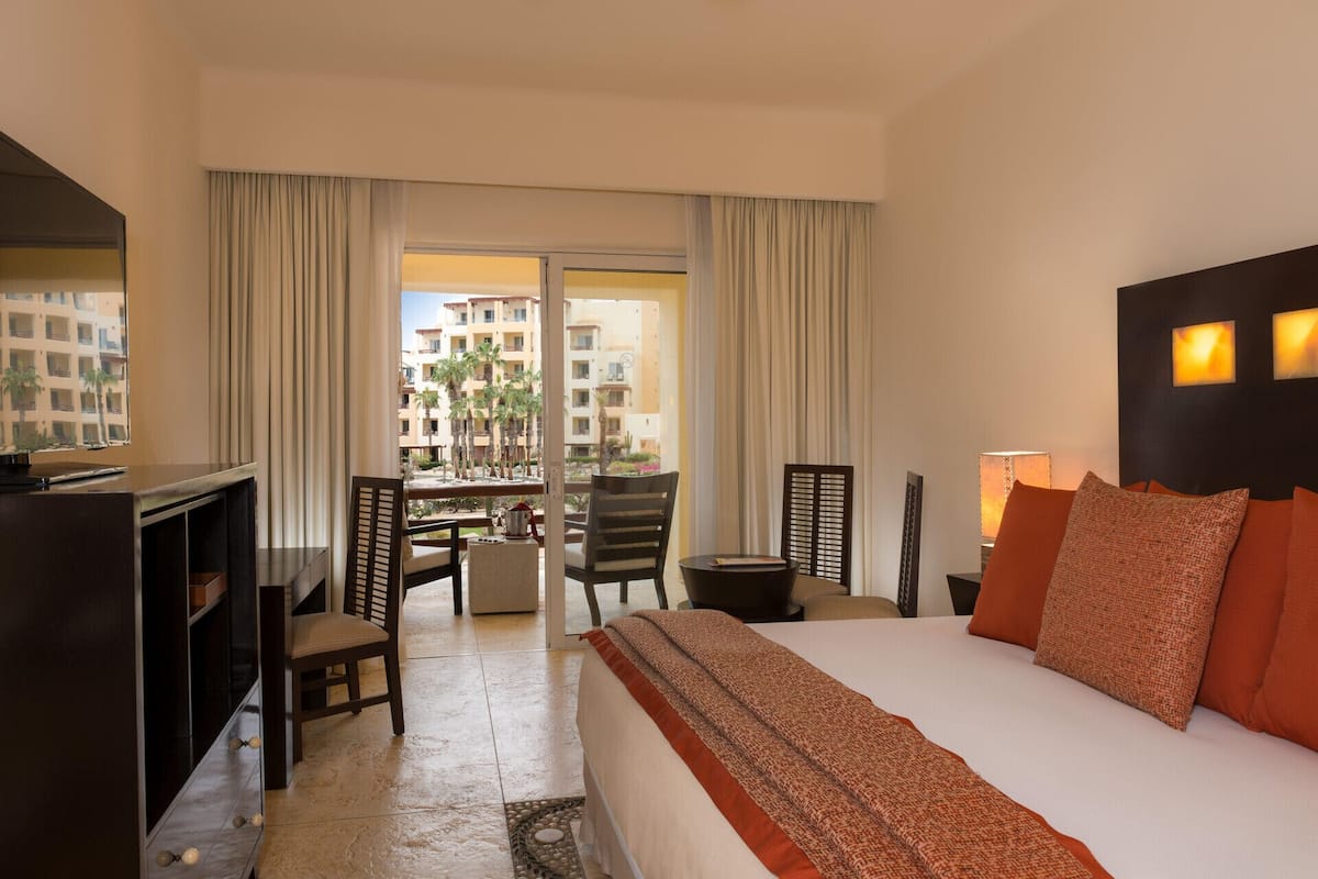 Habitación de hotel con cama, escritorio, silla, lámpara y vista a un edificio con palmeras.