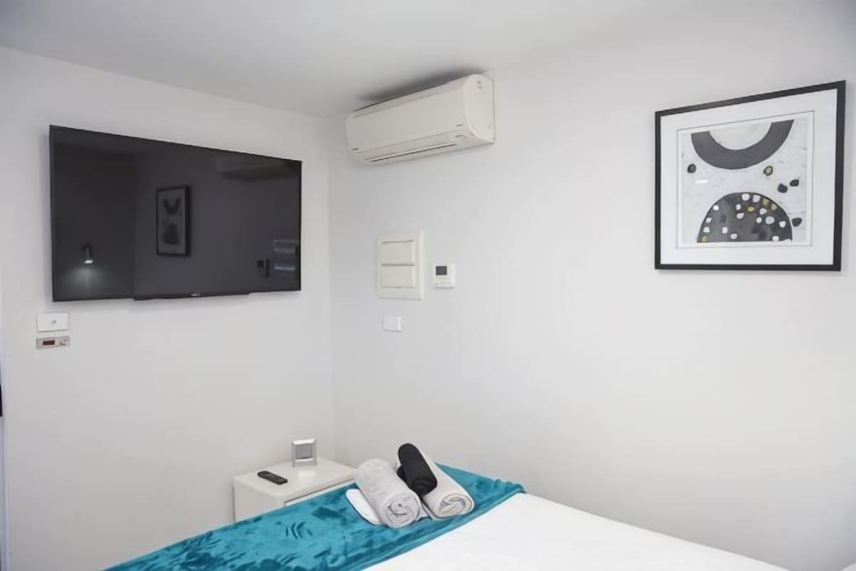 A hotel room with a bed, a TV, an air conditioner, a framed picture, and a bedside table with a phone and a cup.