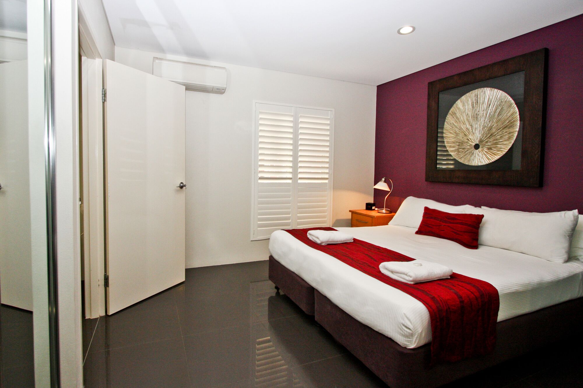 A hotel room with a bed, a nightstand, a lamp, a window with blinds, and a door.