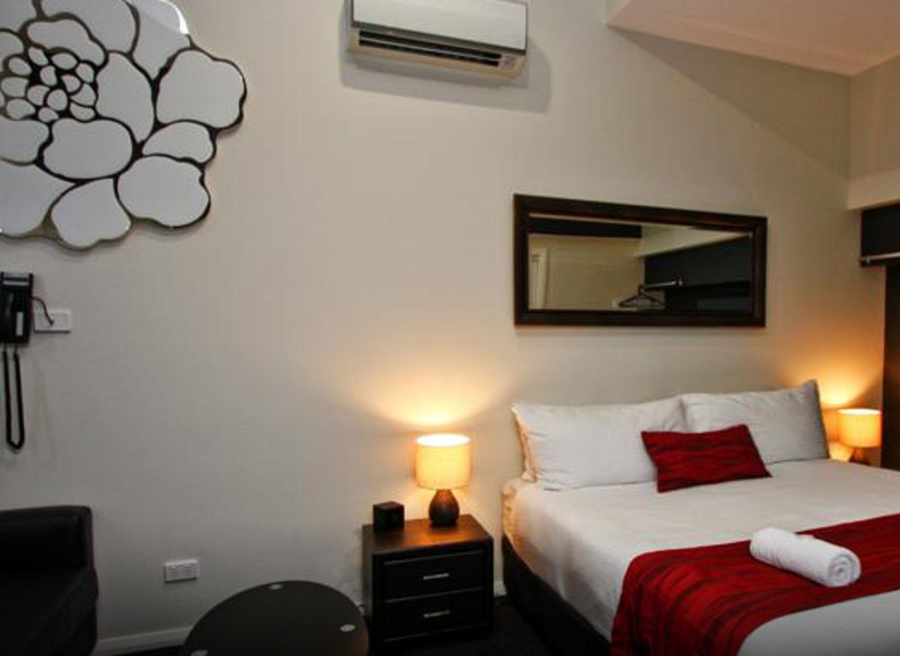 A hotel room with a bed, bedside lamps, a mirror, an air conditioning unit, and a wall decoration.