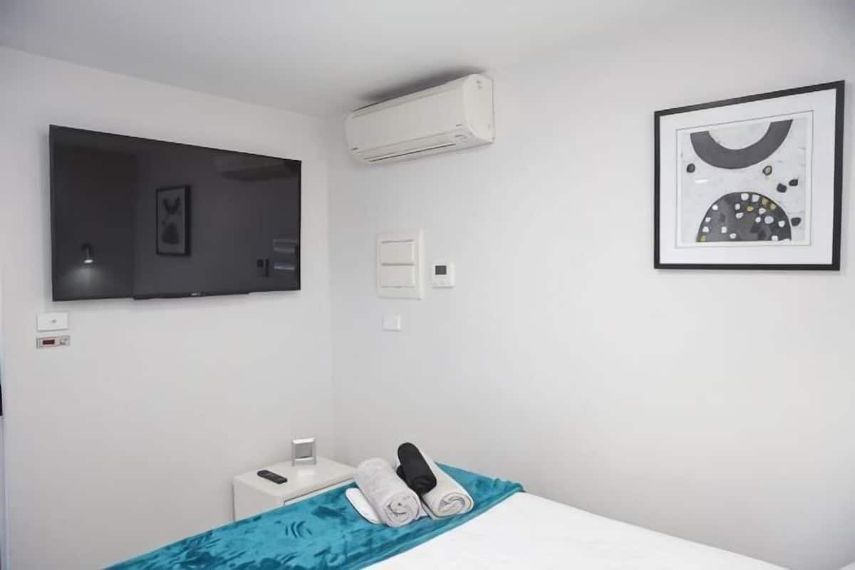 Une chambre d’hôtel équipée d’un lit, d’une télévision, d’un climatiseur, d’un tableau encadré et d’une table de chevet avec un téléphone et une tasse.