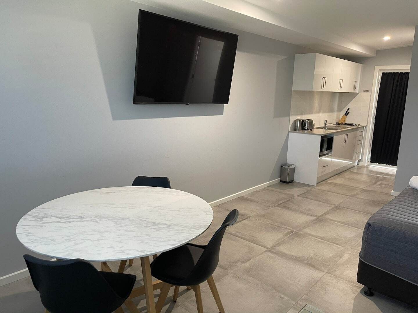 A compact living space with a round marble table, black chairs, a flat-screen TV mounted on the wall, a kitchenette, and a bed.