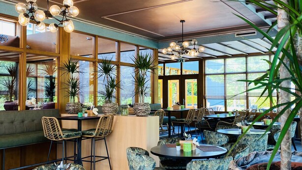 Un restaurante con mesas y sillas redondas, una zona de bar y amplios ventanales que dejan entrar la luz natural.