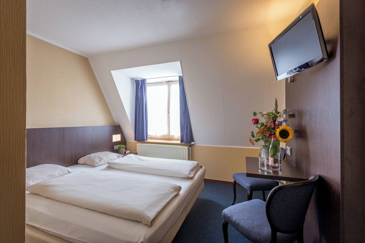 Een hotelkamer met twee bedden, een bureau met stoel, een aan de muur bevestigde televisie en een raam met gordijnen.