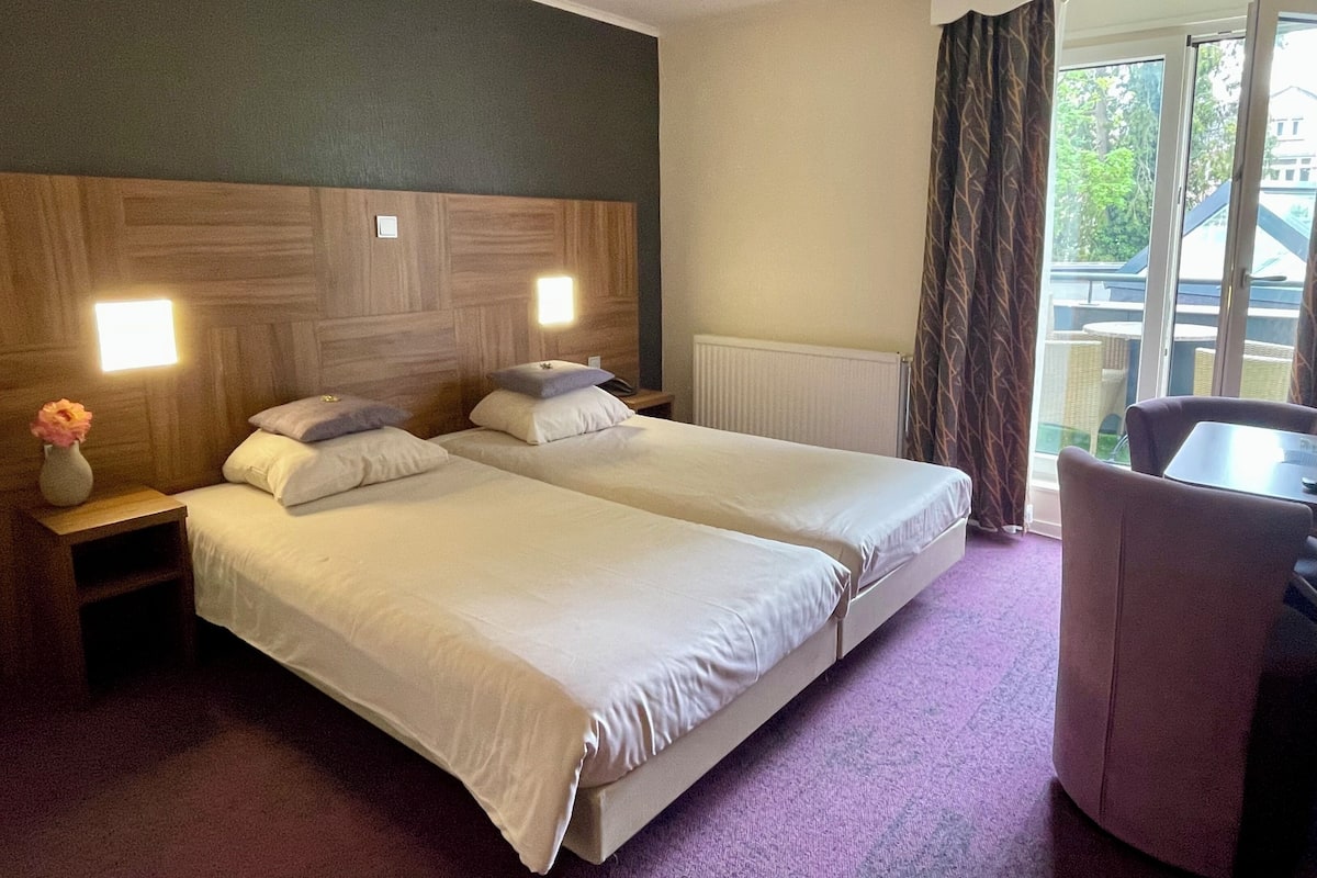 Een hotelkamer met twee bedden, een tafeltje, een stoel en een raam met gordijnen.
