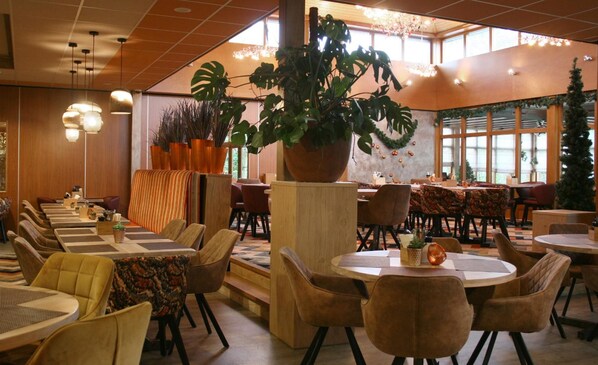 A restaurant with multiple tables and chairs, large potted plants, and a high ceiling with hanging lights.